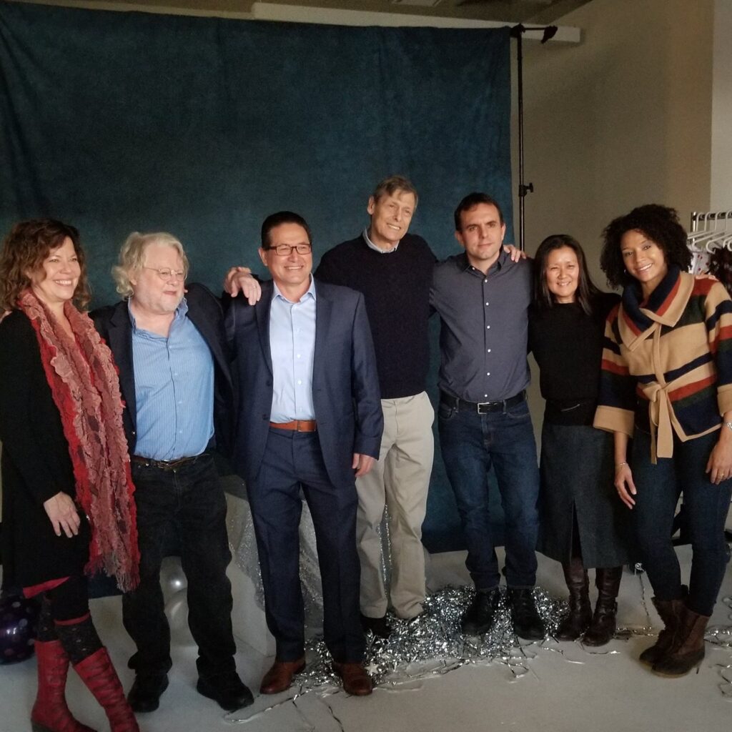 Seven faculty members stand in a photo studio side by side. A backdrop and a rack of clothing sits in the background.