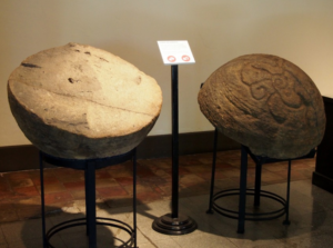 Two halves of a tan, stone sphere are on display. The one on the right is shown with its cut face up, while the one on the left is shown with its cut face down. The one on the left has a swirl pattern engraved in it. In between the halves is a pedestal with a white sign that cannot be read from this image. 