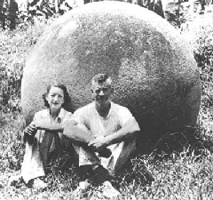 A black and white image of two people, a woman on the left and man on the right, sitting cross legged in front of a stone sphere that is about twice as tall as them in their seated position.