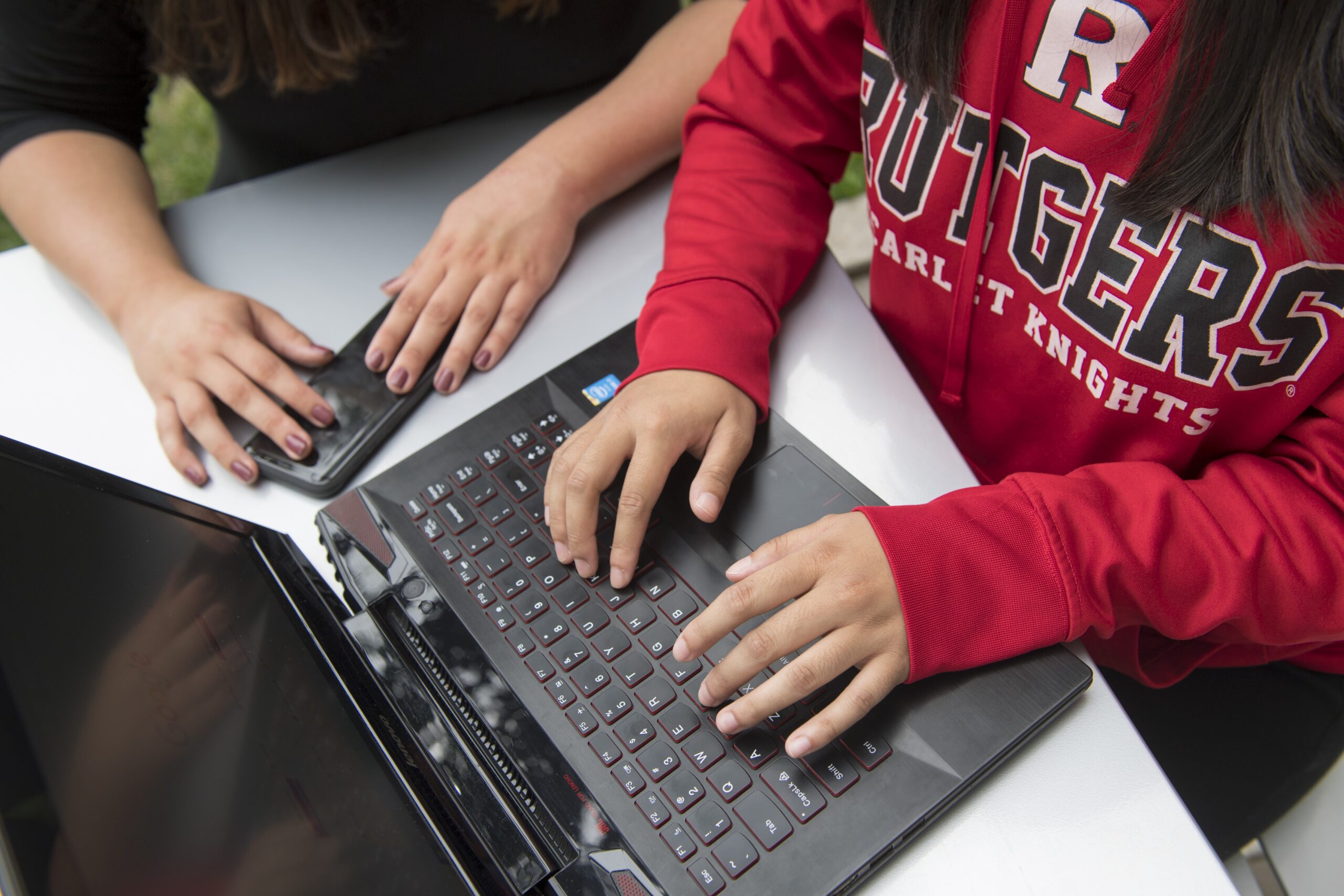A person in a Rutgers sweatshirt typing on a laptop while someone sits next to them, holding a phone