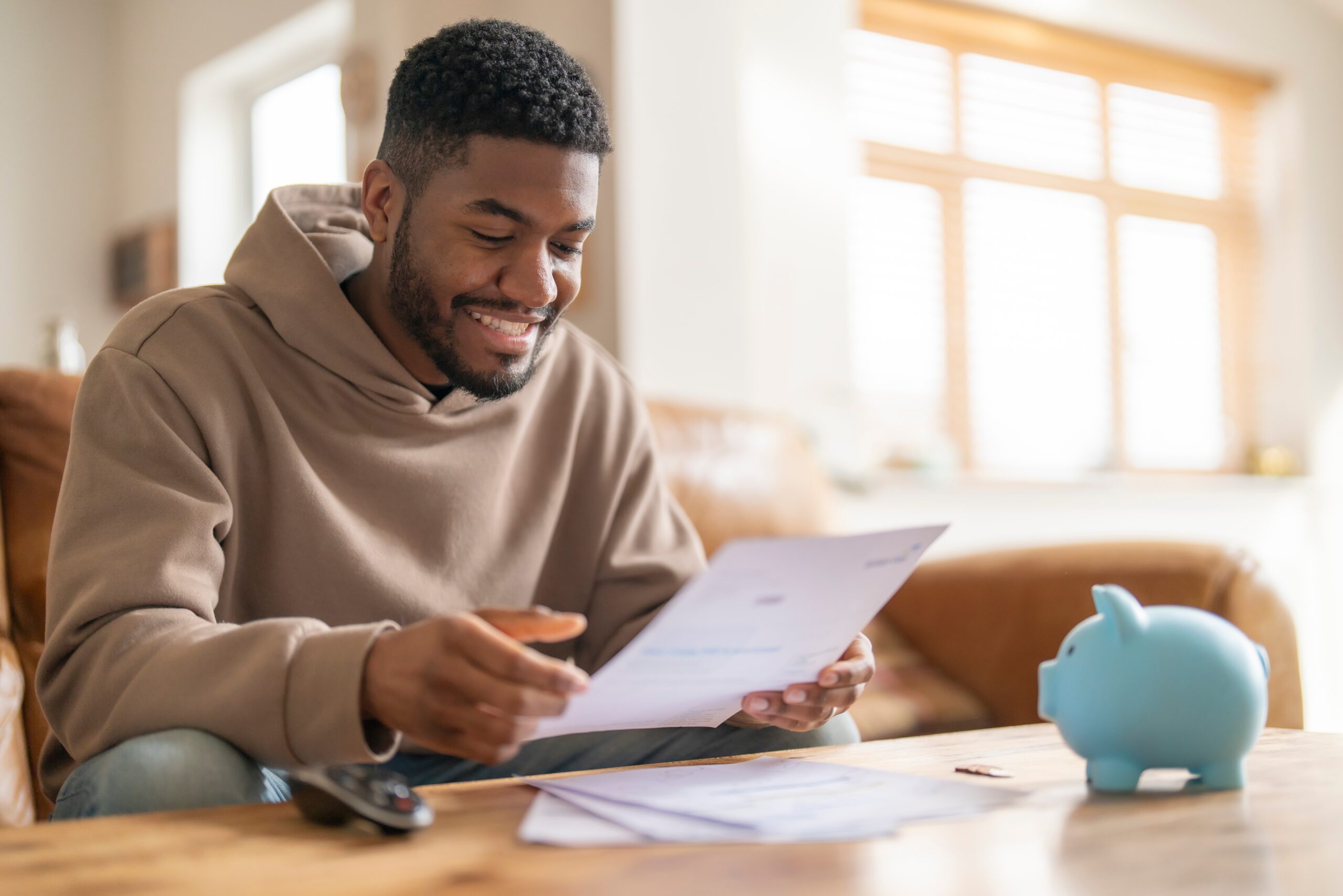 A man smiles as he reviews his finances, papers in hand, with a piggy bank nearby.