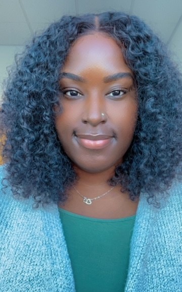 A young woman with curly black hair wearing a green top and a light grey cardigan, smiling in a light background