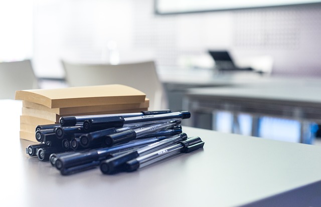 Close-up of a stack of manila sticky notes next to a cluster of black ink pens on a blurred desk in a modern office setting