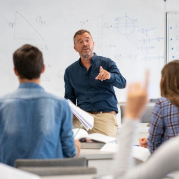 A male teacher in a classroom pointing while engaging with students. There's a whiteboard displaying geometric diagrams in the background.