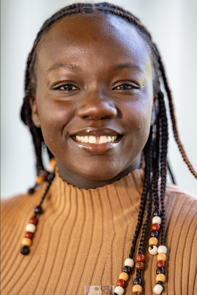 A smiling person with braided hair with colorful beads wearing a brown turtleneck sweater.