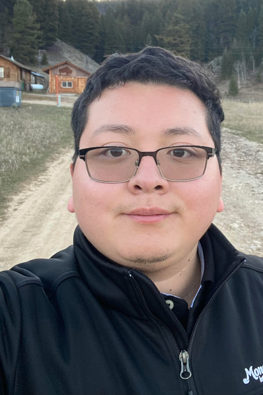 A young person wearing glasses and a black jacket standing on a dirt path with buildings and trees in the background.