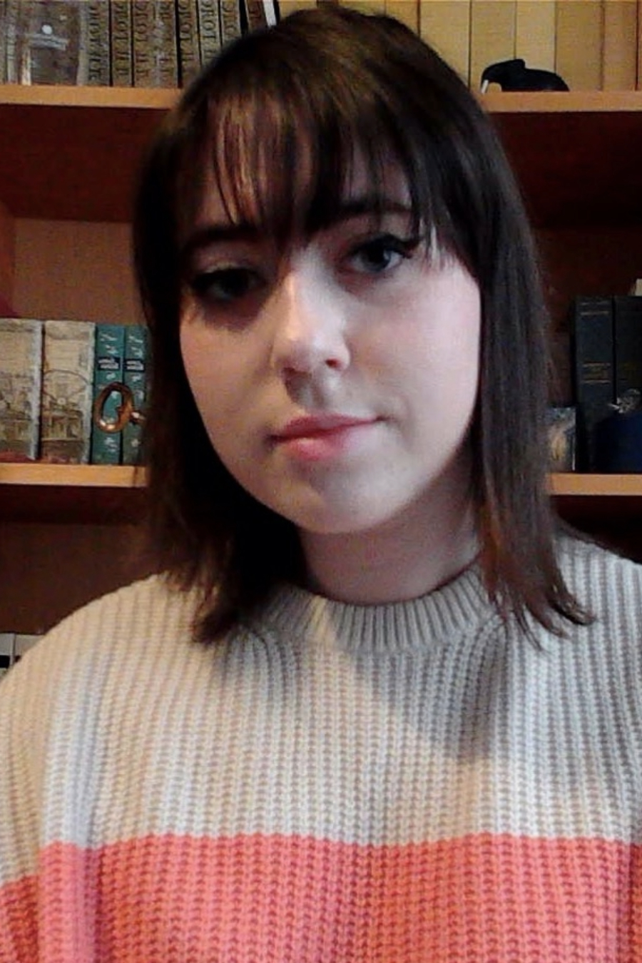 A young woman with dark brown hair and bangs sits in front of a bookshelf, wearing a cream and pink striped sweater