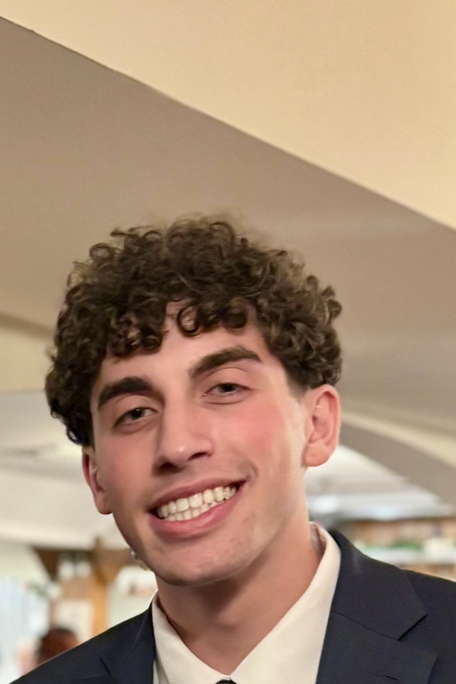 A young man with curly hair smiles broadly while wearing a suit.