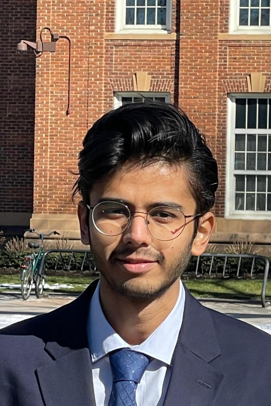 A young man with glasses and dark hair wearing a suit, standing in front of a brick building.