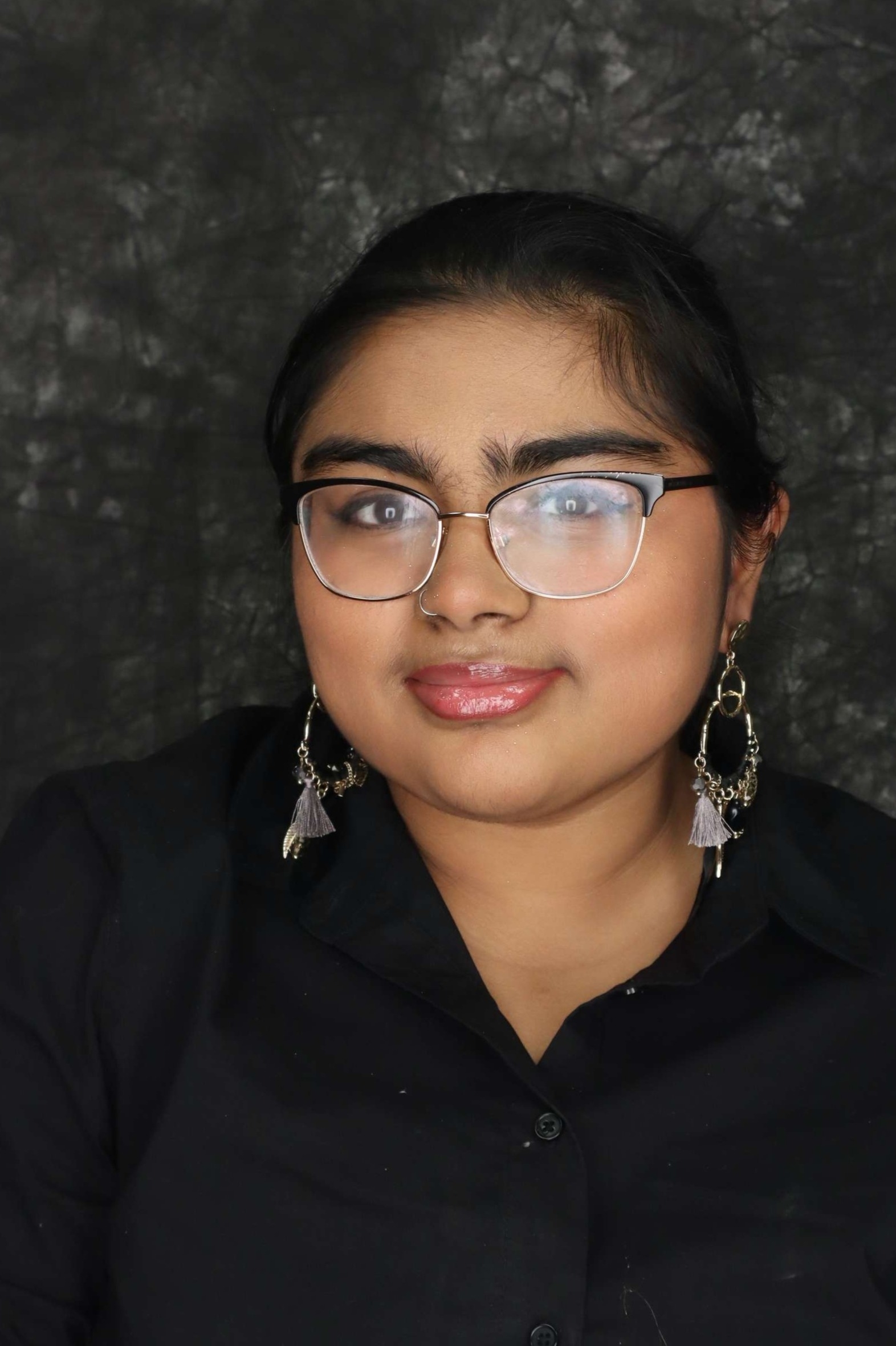 A young woman with dark hair wearing glasses and long earrings against a textured dark background.