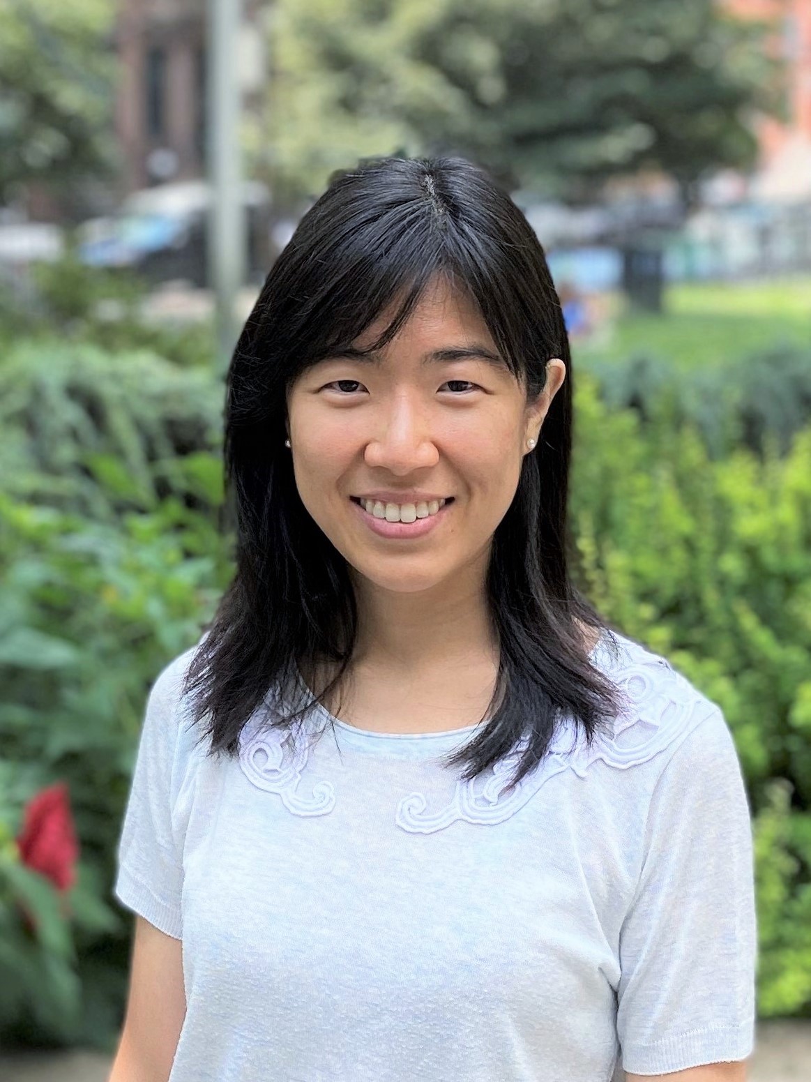 a woman with medium-length black hair wearing a white top, standing outdoors with a blurred garden background.