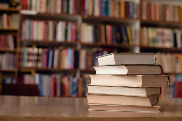 A stack of books sits on a wooden table in a library, with shelves of books blurred in the background.