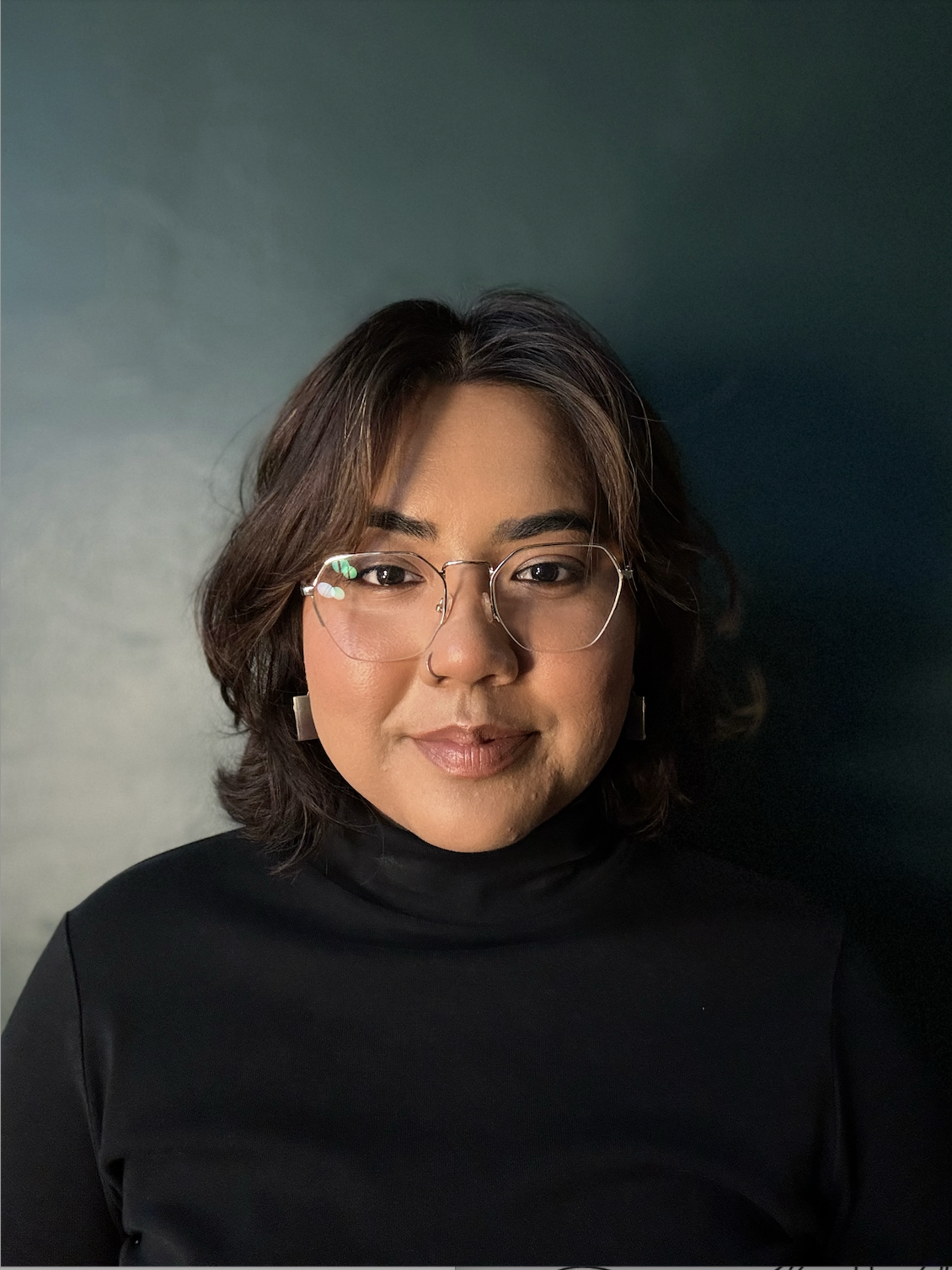 A woman wearing glasses and a black turtleneck poses in front of a dark green background.