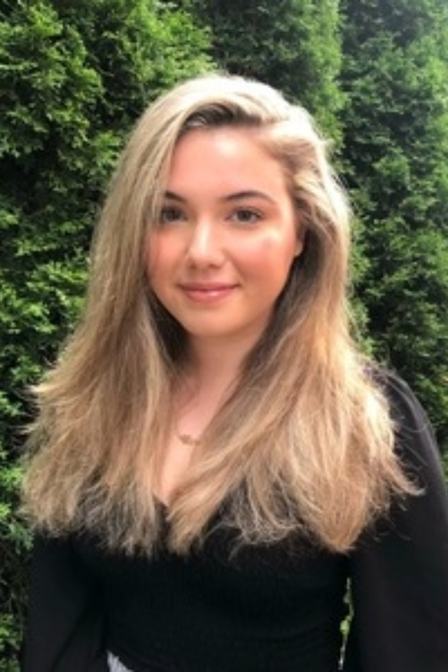 A young woman with long, light brown hair stands in front of tall green bushes, wearing a black top and smiling softly at the camera.