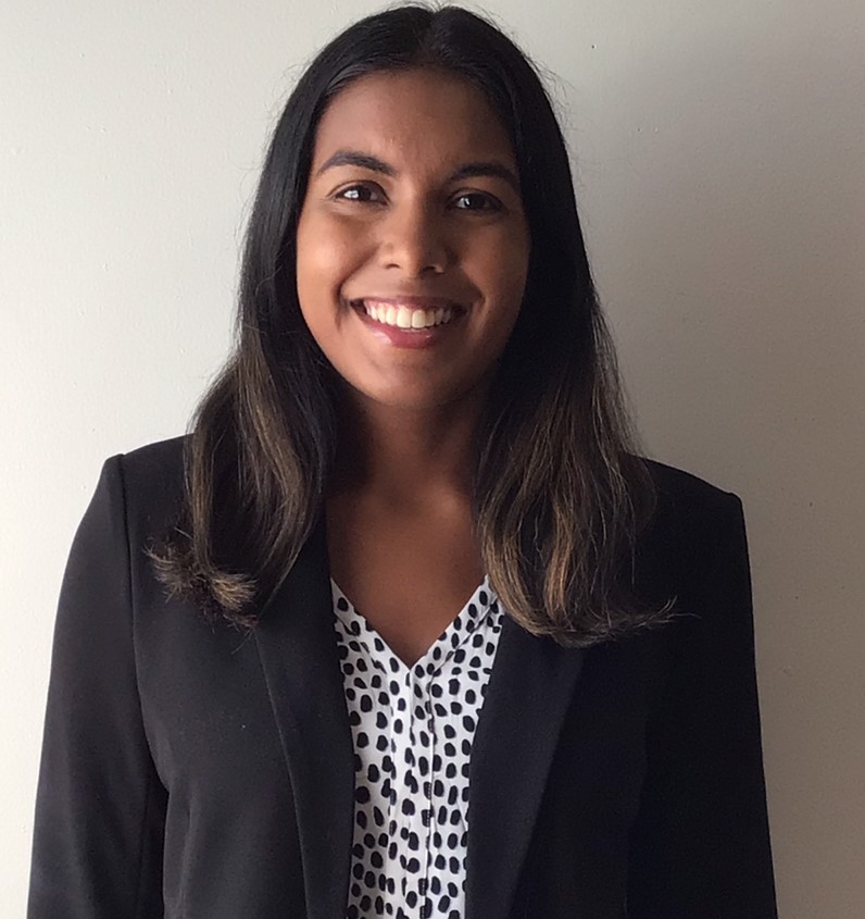 A woman wearing a black blazer and patterned blouse smiles while standing in front of a plain light-colored wall.