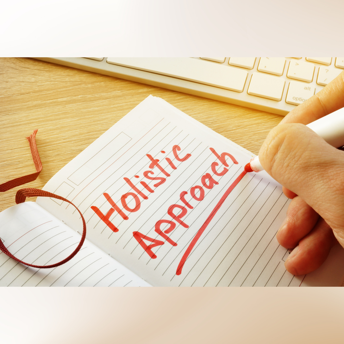 A hand writes the words "Holistic Approach" in red ink on a lined notebook next to a computer keyboard.