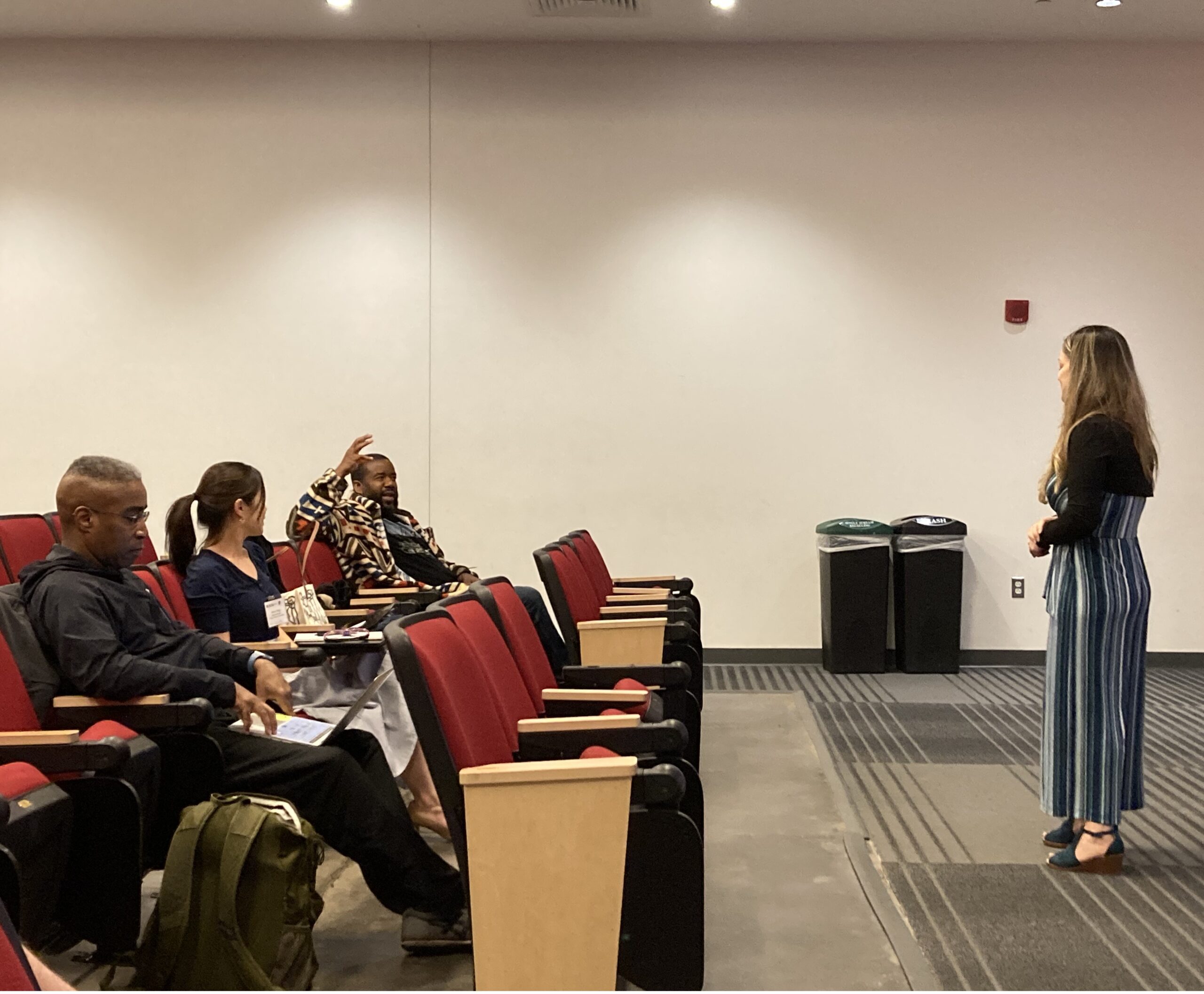 This image shows a lecture-style room with several participants sitting in the first two rows, in discussion with the facilitator, who is standing in the front of the room.