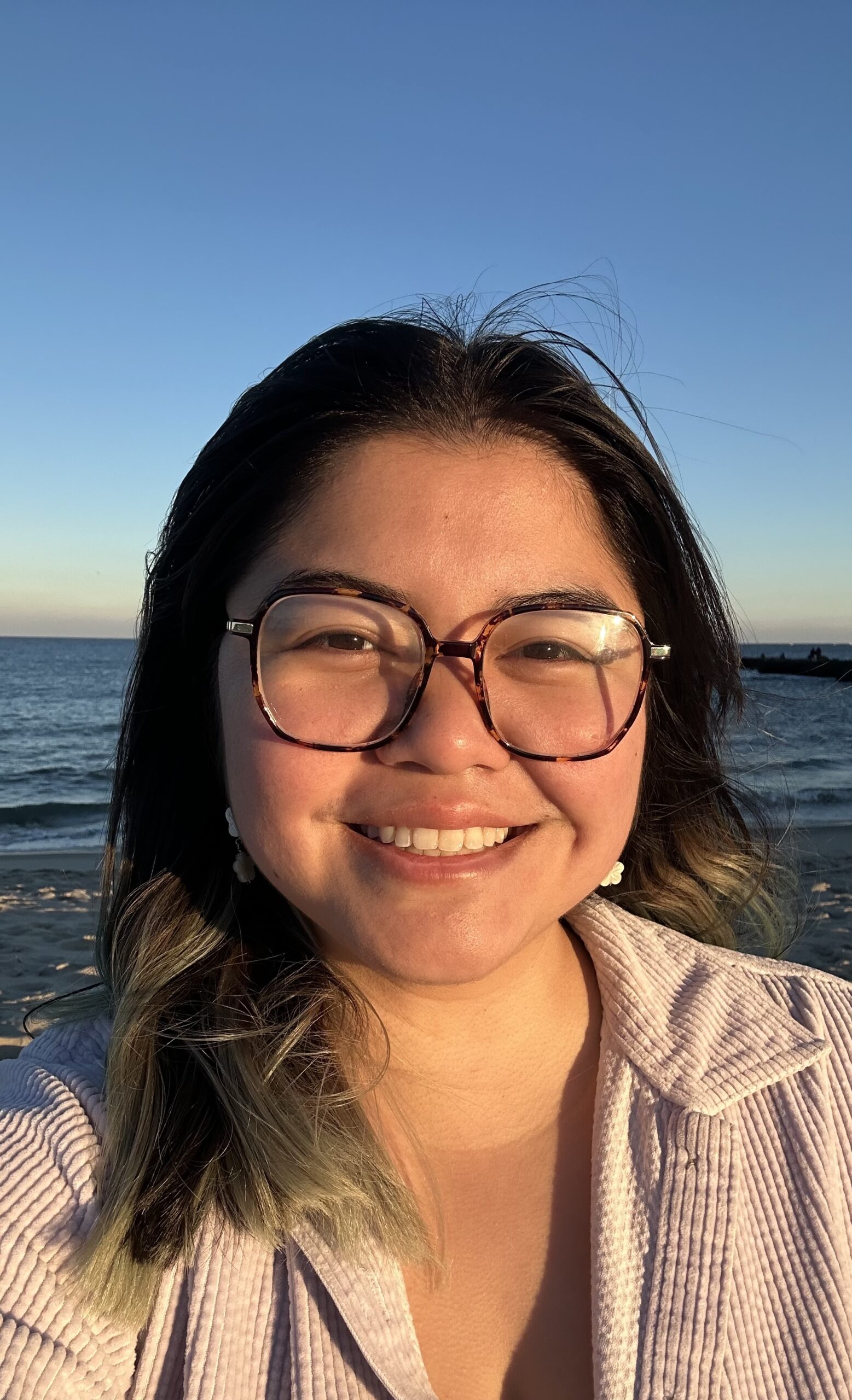 A headshot of a young woman wearing glasses and smiling. Behind her is a blue sky and the ocean.
