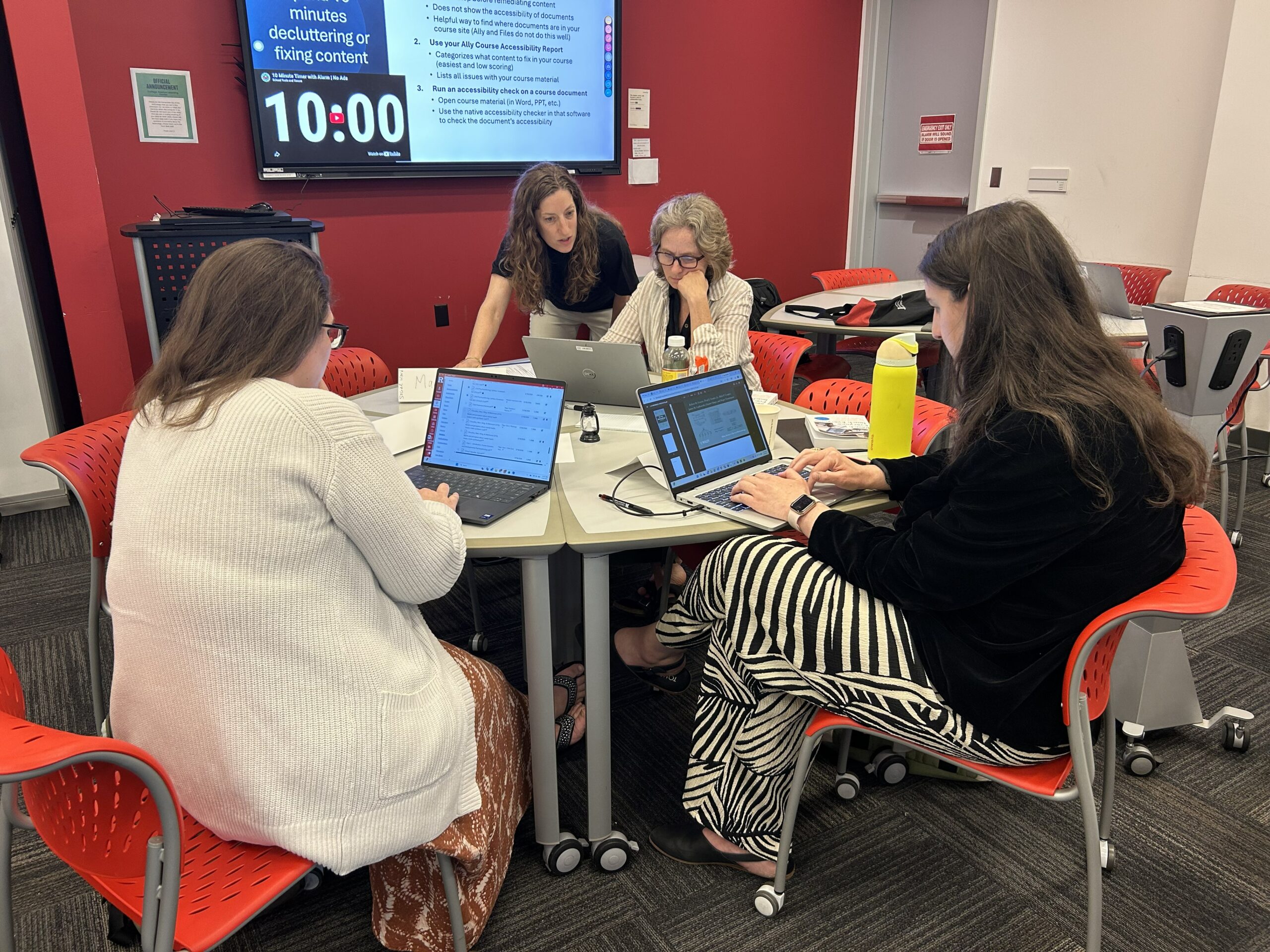Four people seated around a small table in a classroom with red and white walls, each working on a laptop. A large screen on the wall displays a slide titled ten minutes decluttering or fixing content, with bullet points about accessibility tasks and a visible ten‑minute timer.