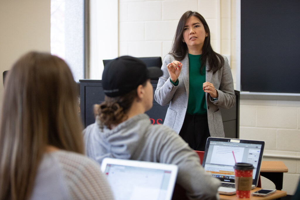 Instructor in a gray blazer gestures while speaking to two students seated at desks with open laptops. A blackboard is behind the instructor, and a window is to the left. One laptop has a red coffee cup beside it.