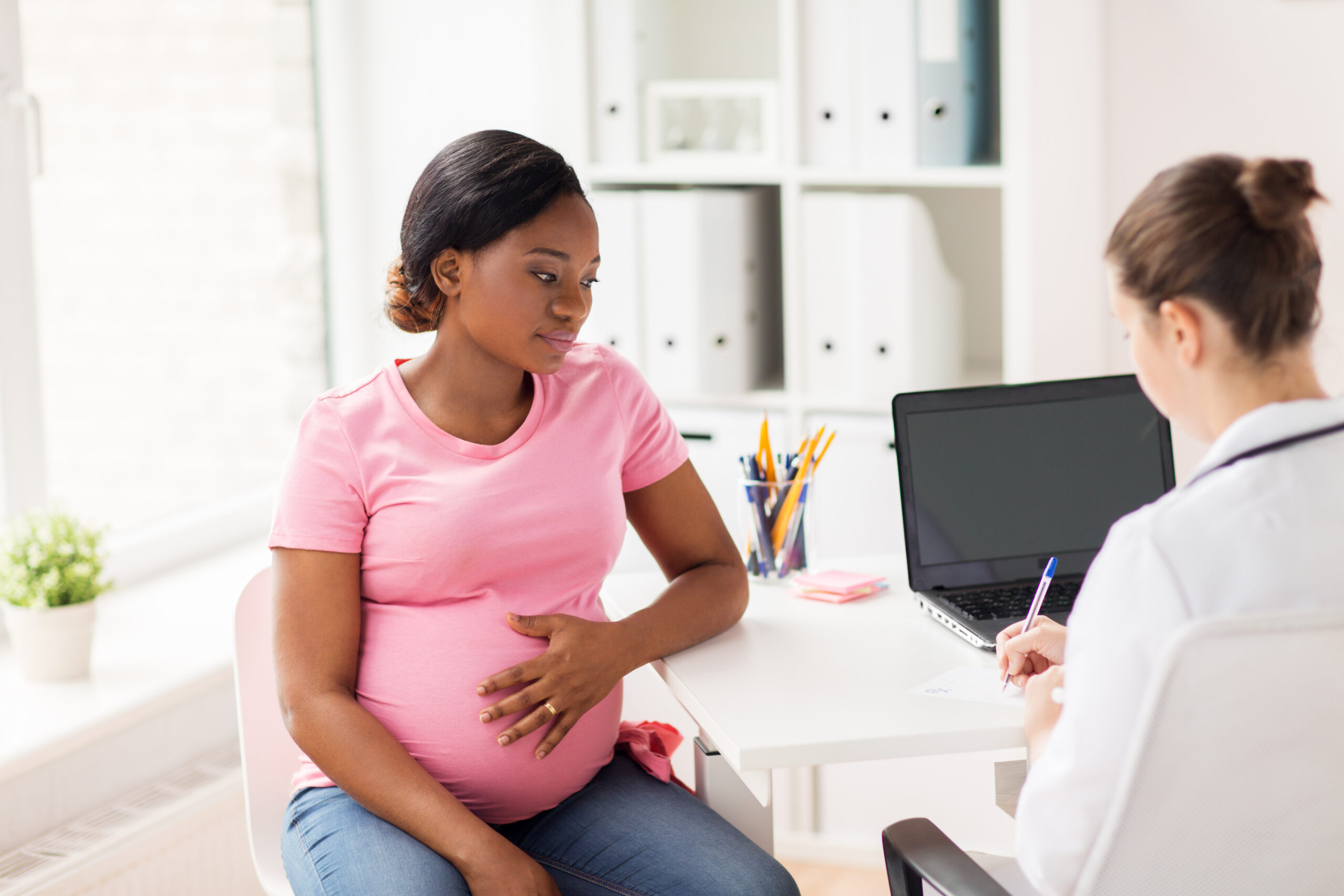 A pregnant Black woman in a pink shirt sits in front of a white female clinician with a laptop