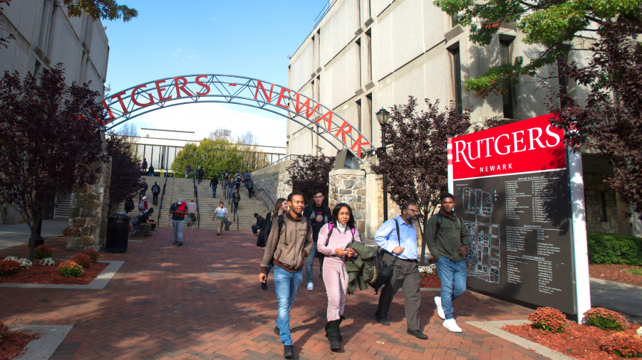 People walking near the Rutgers-Newark arch, staircase, and campus map. Conklin and Boyden Hall are in the background along with several trees.