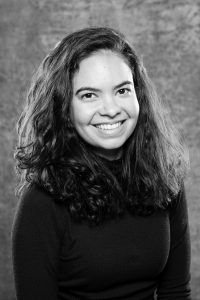 Headshot of a person with shoulder length hair in a black sweater sits in front of a grey marbled backdrop.