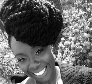 Headshot of a person with an exquisitely braided head of hair smiling in front of some plants in the background.