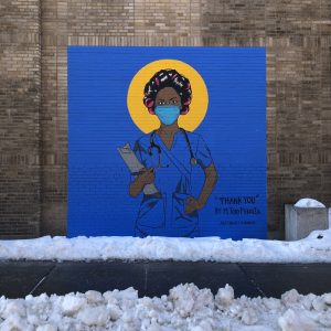 A mural of a nurse is painted on a grey brick wall in front of a sidewalk cleared of the snow on the ground. The black nurse is in blue scrubs matching the background. Her hair is in curlers and she has a mask on. Behind her head is a yellow circle like a halo. The piece is titled "Thank you" and signed M. Tony Peralta.