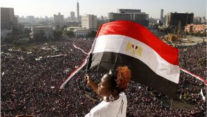 A woman waves an Egyptian flag in the foreground. below is a crowd of thousands holding signs and banners. A city sits beyond the crowd.