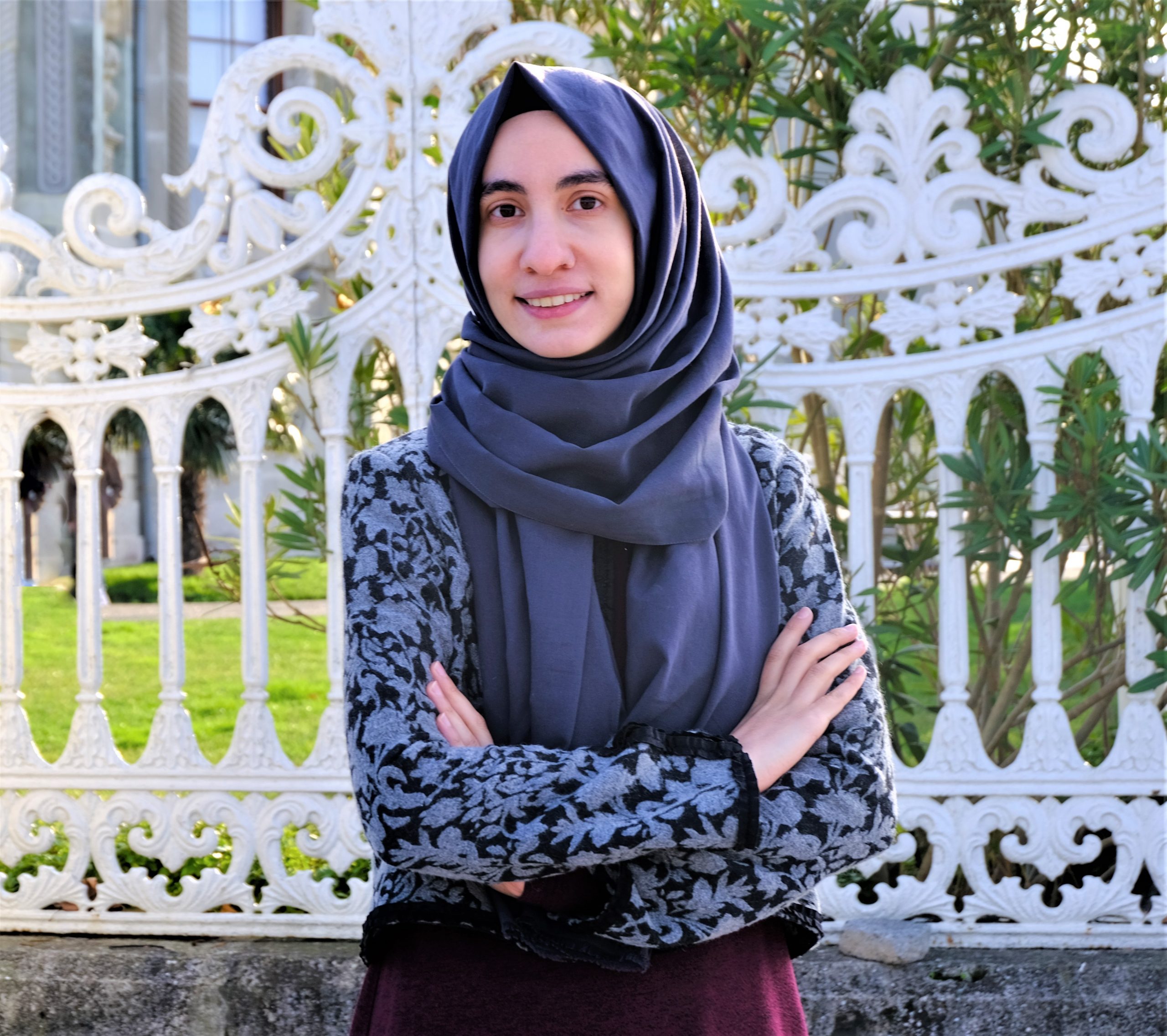 Headshot of Fatma Zisan Tokac standing in front of a white sculpted fence. Greenery and a building stand blurry in the background.