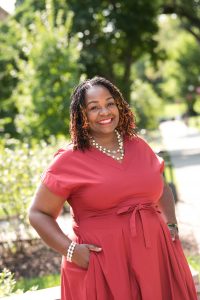 Headshot of Hyacinth Miller. She stands in the foreground in a coral dress. Trees and a path sit in the background.