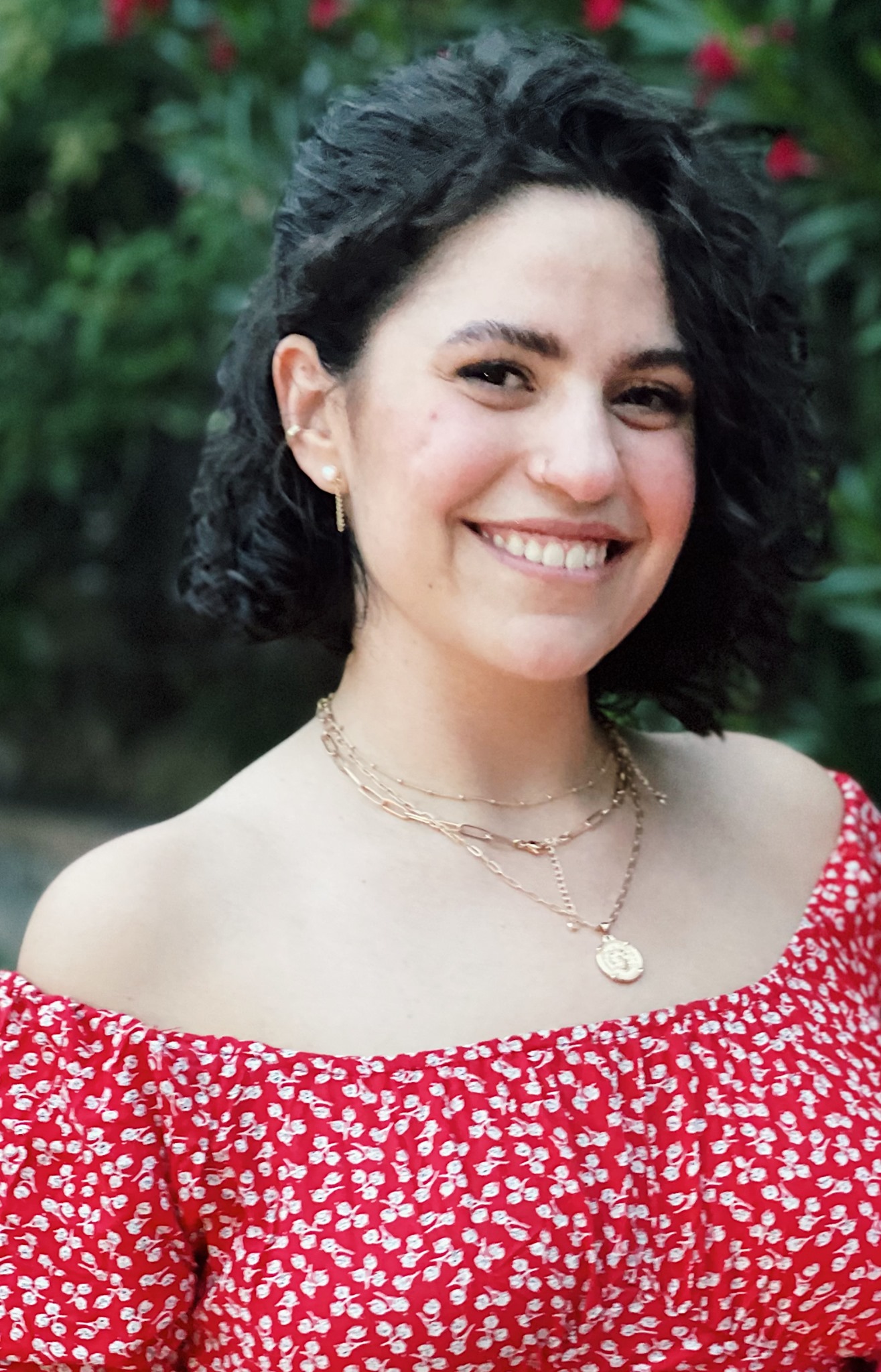 Headshot of Ana Sondoval. Greenery sits in the blurry background. Sandoval is wearing a red and white floral top and some gold necklaces.