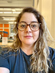 Headshot of a Cintia Martins Freitas. She wears glasses and a blue V-neck t-shirt and looks into the camera.