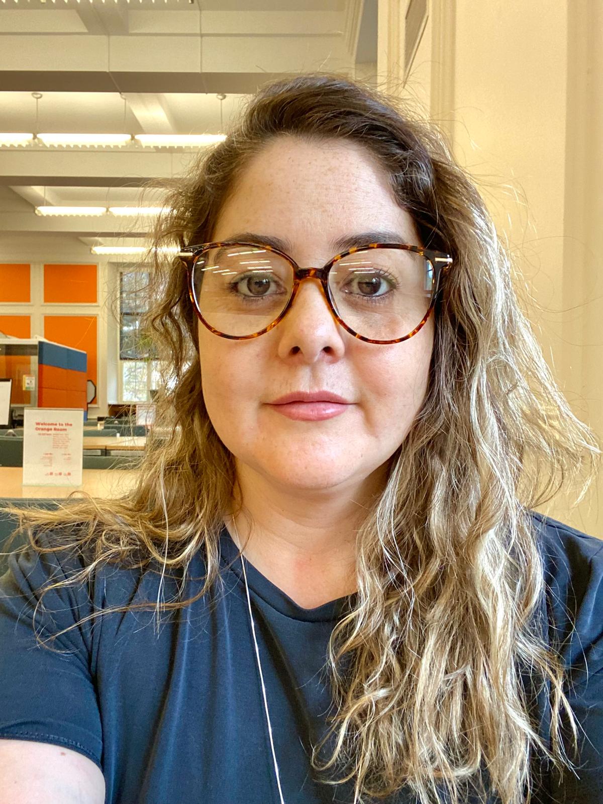 Headshot of a Cintia Martins Freitas. She wears glasses and a blue V-neck t-shirt and looks into the camera.