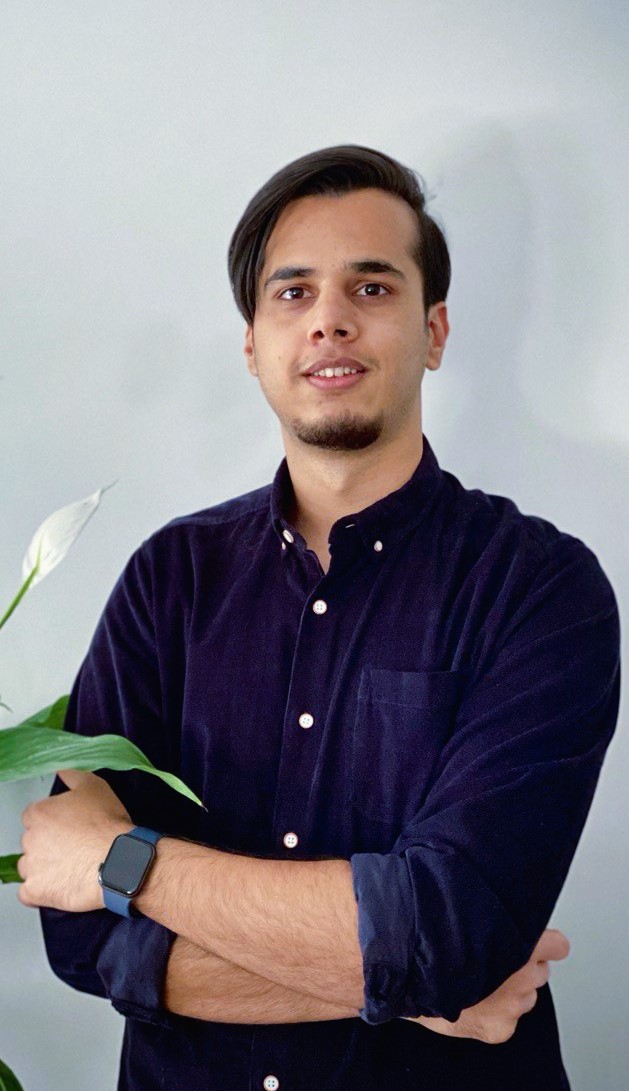 Headshot of Mustafa Hoshmand. A person with dark hair in a dark blue button up looks at the camera. A lily peeks into the frame in the foreground.