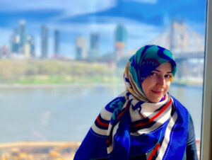 Person in a colorful headscarf with a blurry cityscape and body of water in the background.