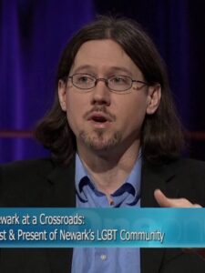 Screengrab of Whitney Strub being interviewed on a television show. A banner on the bottom of the photo reads "Newark at Crossroads: Past & Present of Newark's LGBT Community"