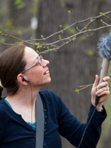 Rachel Mundy in a wooded area holding a microphone while looking toward a tree out of frame.