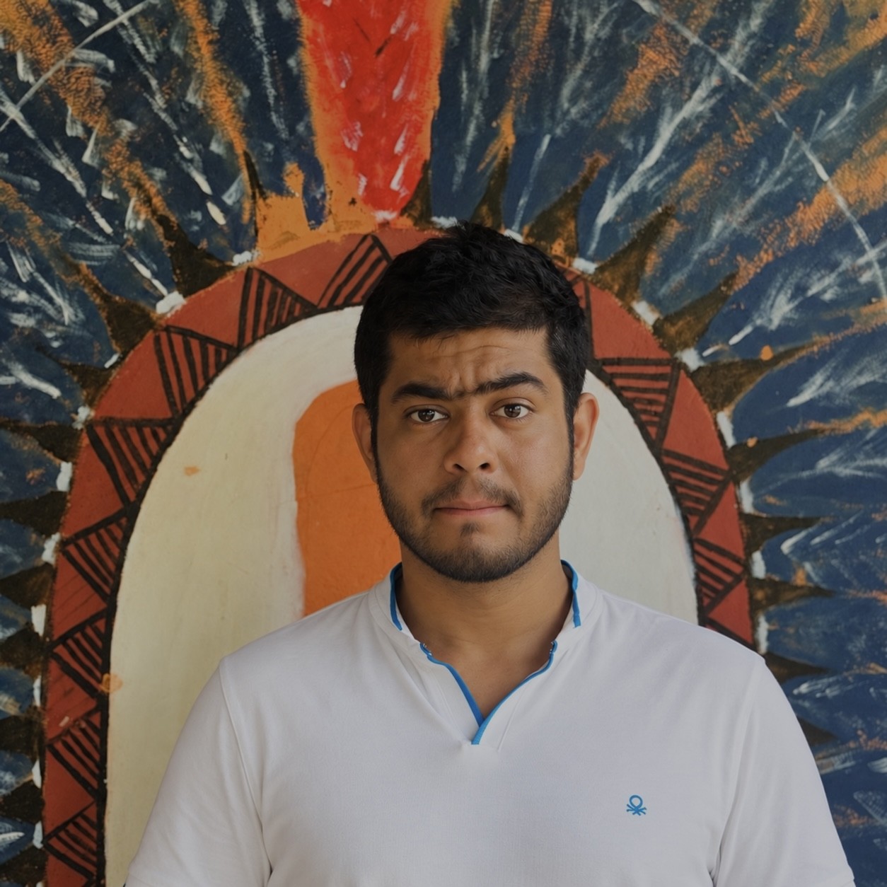 Portrait of a person wearing a white collared shirt, standing centered in front of a colorful mural. The mural features a large circular design in orange, white, black, and blue, with radiating brushstroke patterns that create a halo-like effect behind the person's head. The person is facing the camera against the vibrant, abstract background.