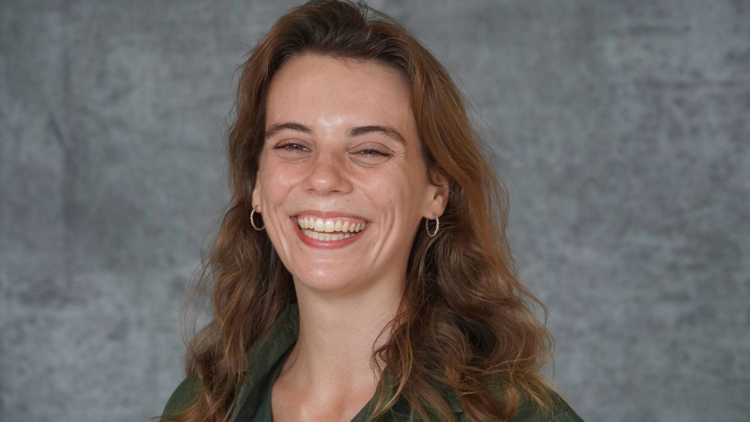 Professional headshot of a person with shoulder-length wavy brown hair wearing small hoop earrings and a dark green collared shirt. The person is facing the camera with a broad smile against a mottled gray studio backdrop. The image is evenly lit and framed from the shoulders up.