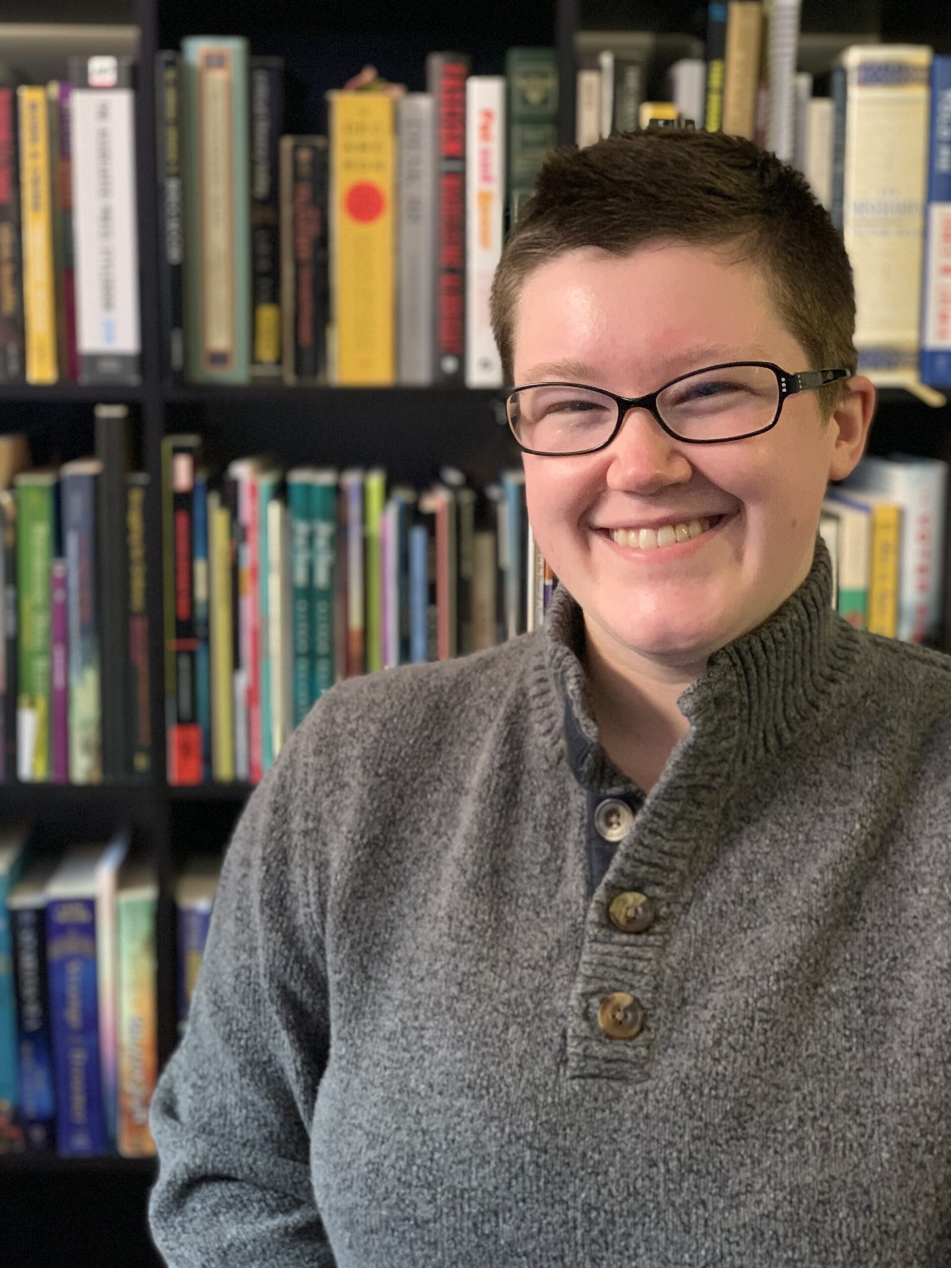 Moira Armstrong, a white person with black glasses and short brown hair wearing a gray turtleneck sweater, stands in front of a bookshelf, smiling.